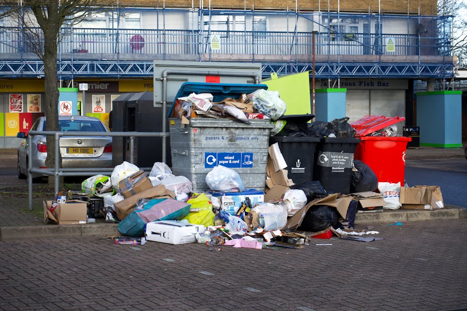 A black wheelie bin labeled 'ST. JOHN'S' positioned on a paved sidewalk at night, filled with various discarded items including cardboard boxes, paper, and miscellaneous rubbish spilling over the top.