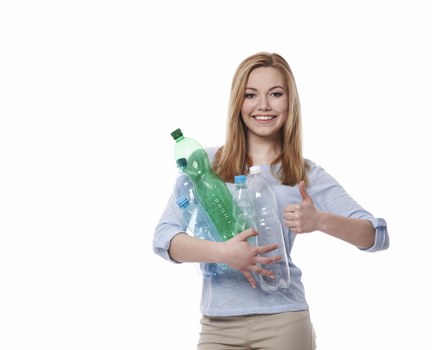A young woman with long, straight, reddish hair and light skin is standing against a plain white background, smiling confidently while holding several clear plastic bottles filled with liquid or empty recycling bottles. She is dressed in a light blue, long-sleeved shirt with rolled-up sleeves and beige trousers. The bottles in her arms vary in size and shape, with some having green or blue caps, and are positioned close to her torso, held securely with one hand supporting them from underneath. She is giving a thumbs-up with her other hand, indicating approval or success. The lighting is even, highlighting the transparent textures and subtle variations in the plastic bottles. Although the scene appears staged for a promotional or environmental message, the setting is minimalistic, emphasizing the bottles and the woman's positive gesture, subtly aligning with rubbish and recycling services in Highgate or the local area served by Flat Clearance Highgate, operating within N6 postcode boundaries.