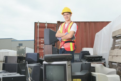 A male worker wearing a yellow safety helmet and a high-visibility vest stands outdoors at a waste collection site, surrounded by various discarded electronic appliances including old computer monitors, televisions, and other electronic waste items. The appliances are spread across the ground and appear to be stacked in a haphazard manner. In the background, there are shipping containers, a warehouse-style building, and an overcast sky. The worker is smiling and has his arms crossed, indicating a professional approach to waste or rubbish removal services. The scene is set in an industrial area, possibly within or near Highgate, with a focus on the collection and disposal of electronic waste, which is a typical task for Flat Clearance Highgate and other rubbish removal specialists in the area.
